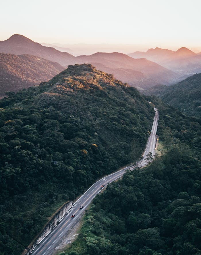 Drone captures a winding road through lush mountains at sunrise, offering stunning scenery.